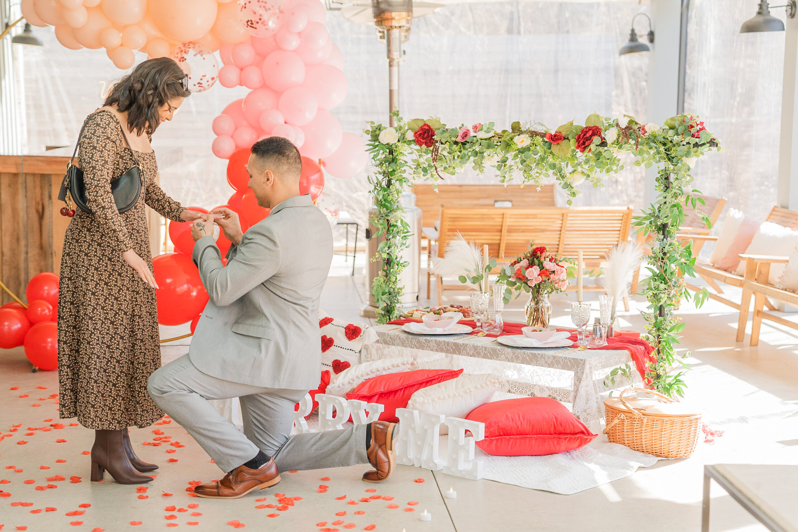 A guy kneeling down putting a ring on the left hand of a woman. The guy wearing a casual suit and the woman is wearing a brown long dress with a dark brown booties and close to them is a picnic table with full of flowers and a charcuterie board and a balloon arch background