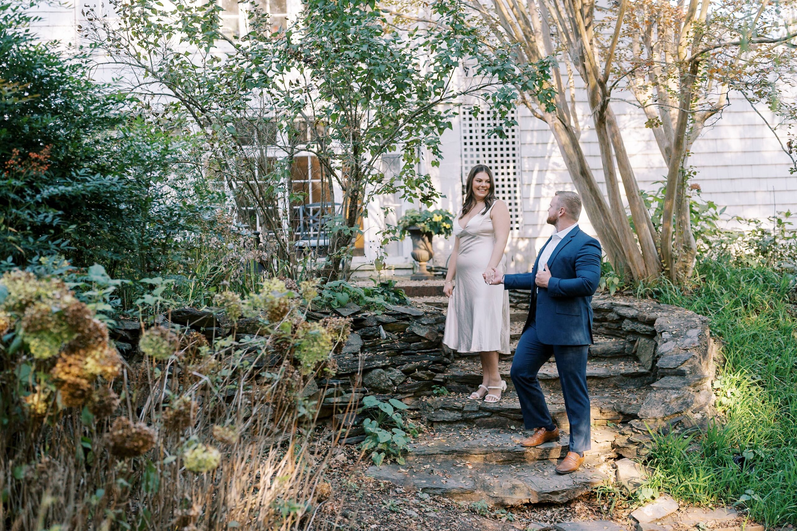 A couple poses for their engagement session in a lush garden, with the woman in an elegant champagne dress and the man in a navy suit. Captured in natural light, the scene exudes romance and timeless charm. Photographed in Charlottesville, VA